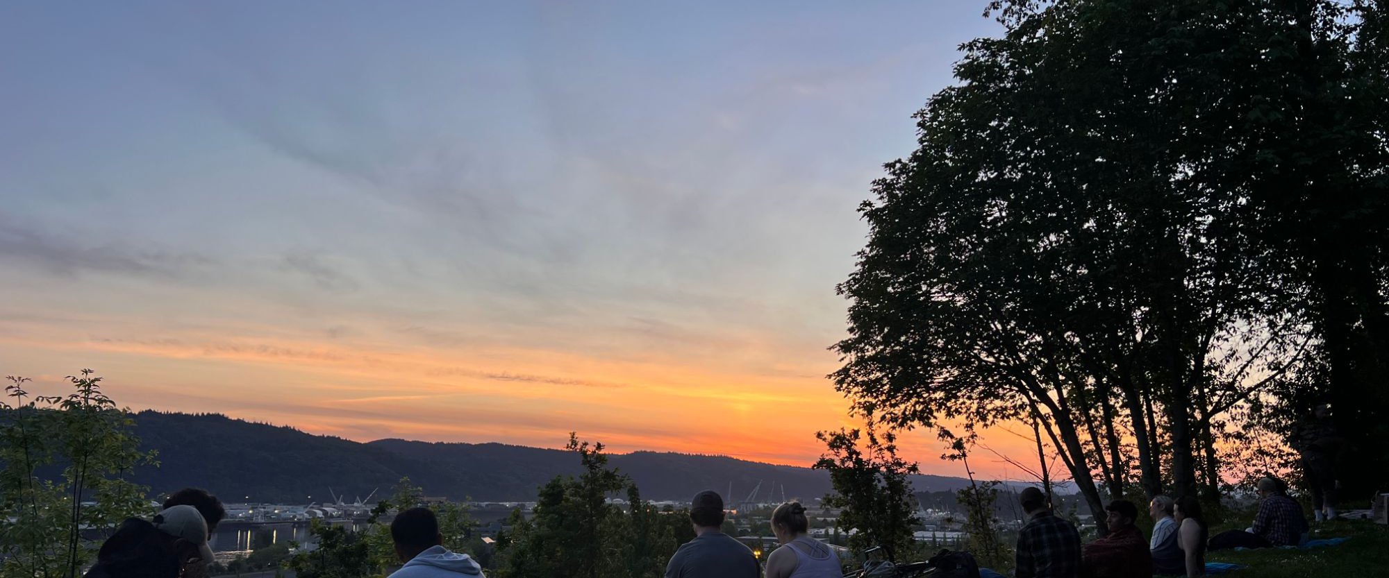 Sunset over the hills and river of Portland, Oregon, with people sitting on the grass